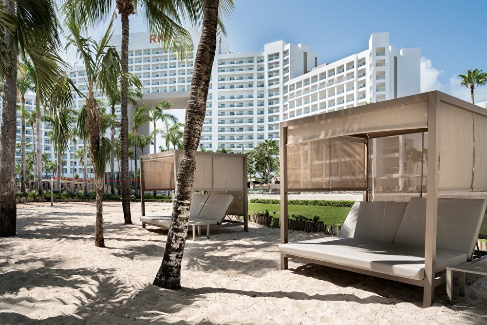 amas balinesas privadas sobre la arena blanca de la playa, rodeadas de palmeras con el imponente edificio del hotel Riu Palace Península al fondo