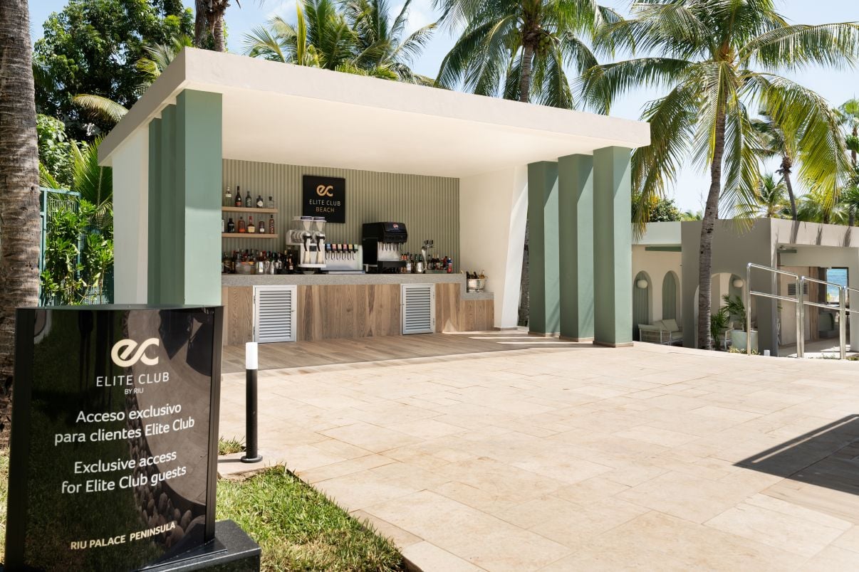 Bar al aire libre «Elite Club Beach» con barra de servicio bajo una estructura moderna verde y blanca, rodeado de palmeras y con un cartel de acceso exclusivo en primer plano
