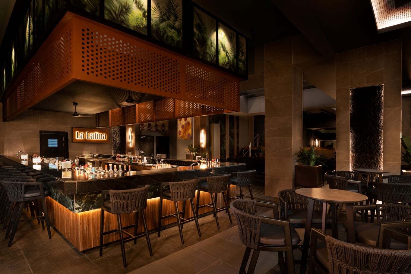 Bar de coctelería «La Latina» con una barra envolvente de mármol, iluminación cálida tenue, taburetes altos de tono oscuro y decoración con plantas tropicales