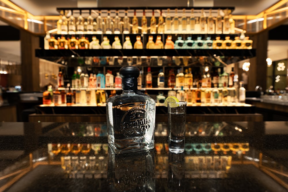 Una botella de tequila Don Julio 70 y un caballito de tequila con escarchado de sal y una rodaja de limón sobre una barra de mármol negro con repisas de licores iluminadas de fondo