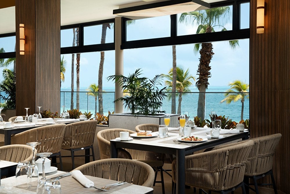 Comedor de restaurante moderno con mesas puestas para el desayuno, sillas de fibra tejida y amplios ventanales con vistas panorámicas al mar Caribe y a las palmeras