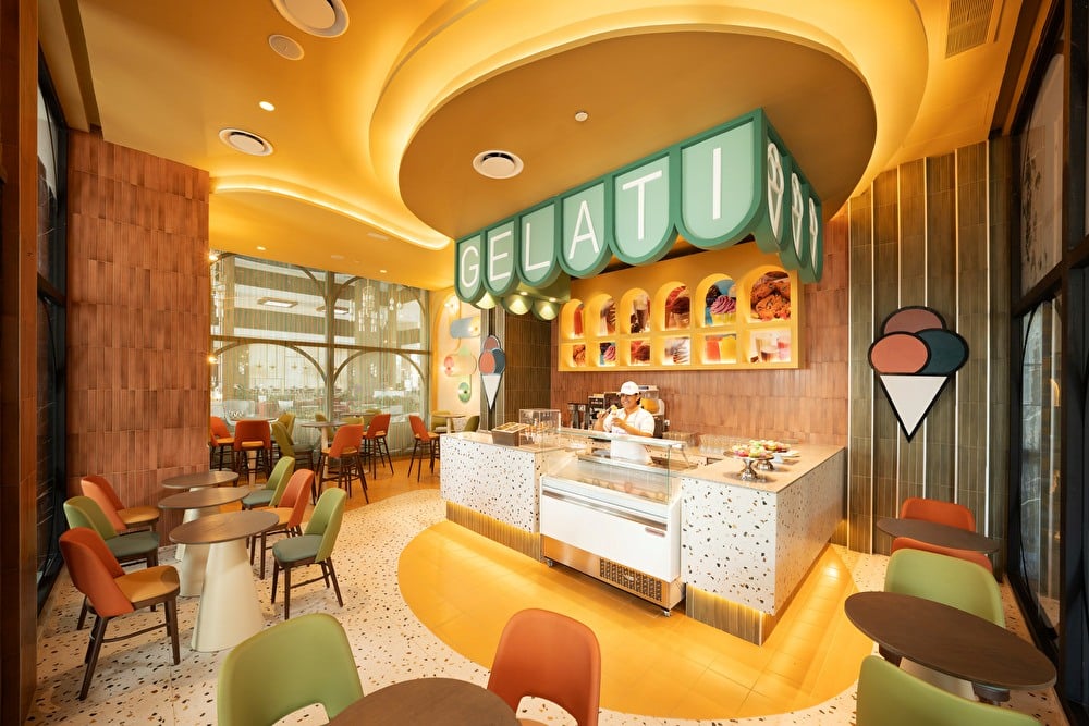 Heladería interior de estilo retro con un mostrador vitrina de helados, letrero luminoso de «Gelati», tonos cálidos y mesas redondas con sillas de colores