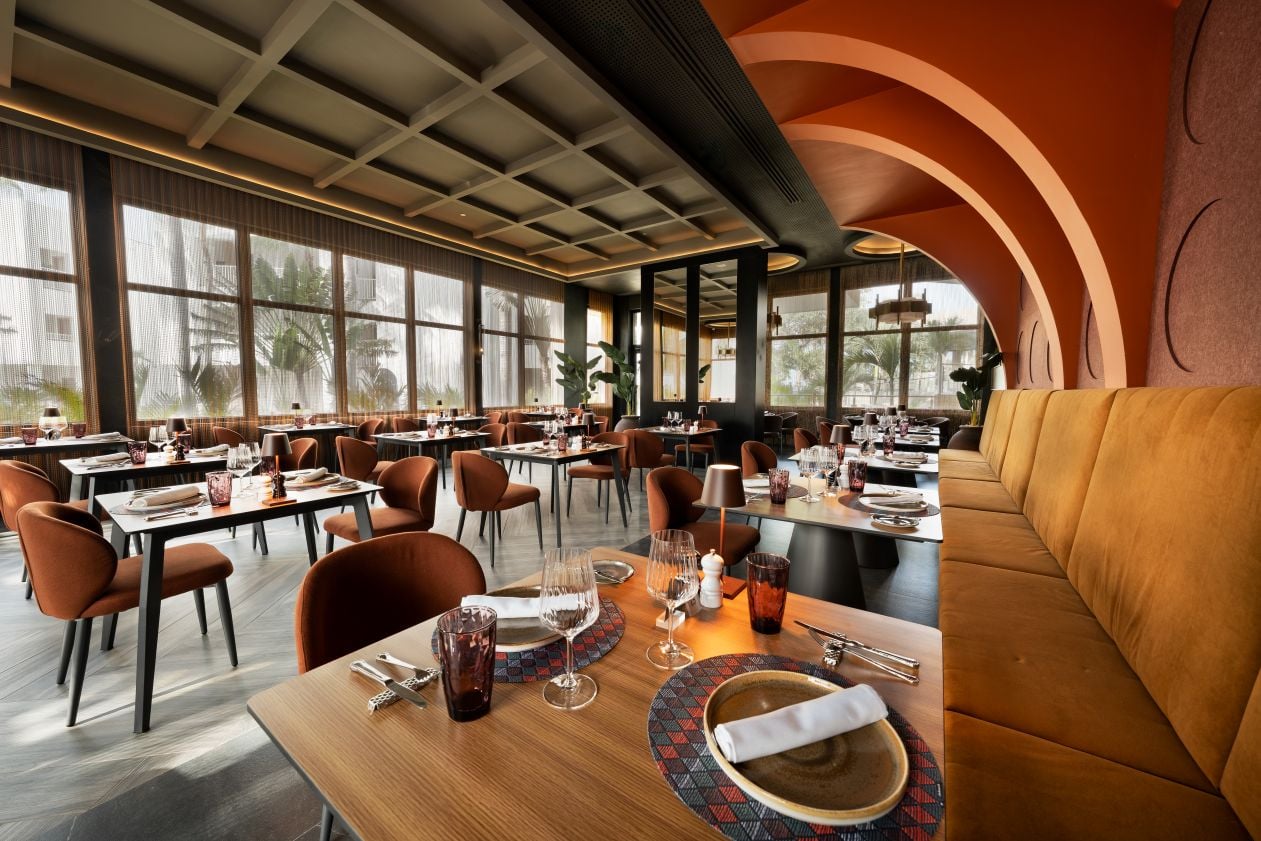 Salón comedor de restaurante elegante con mesas vestidas con copas y vajilla, banquetas acolchadas en tono mostaza, paredes con arcos terracota y ventanales luminosos