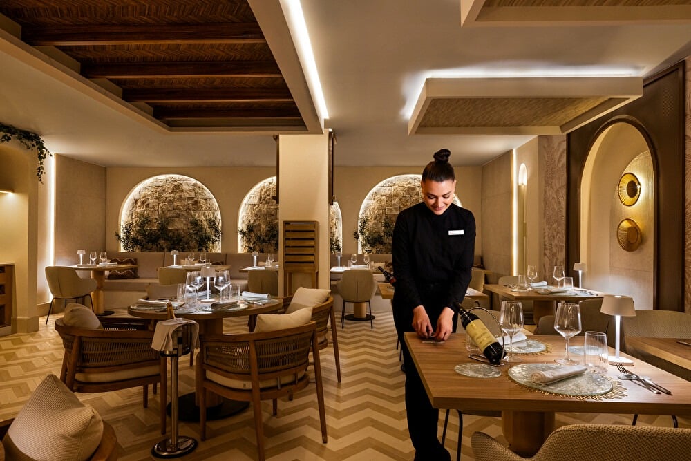 Camarera colocando vino en una mesa de comedor beige con suelo de parquet en el restaurante Krystal del hotel Riu Palace La Mola
