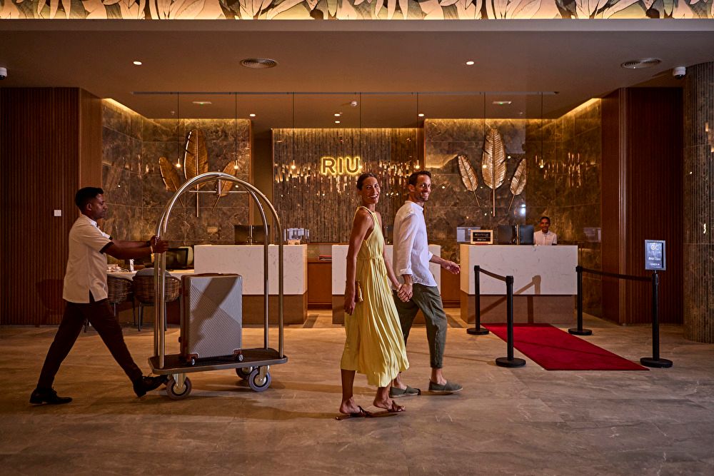 Pareja caminando sonriente por el lobby del hotel mientras un botones lleva sus maletas en un carro de equipaje cerca de la recepción.