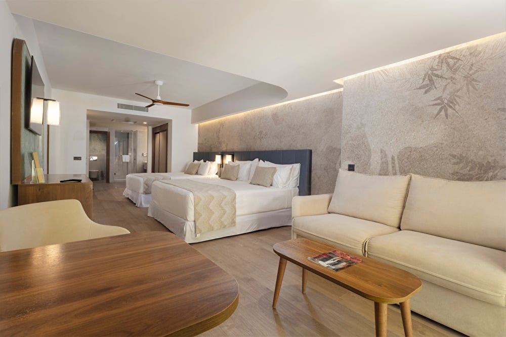 Family Room at Riu Palace Aruba featuring two double beds and a lounge area with a sofa, decorated in cream tones with wooden furniture details.
