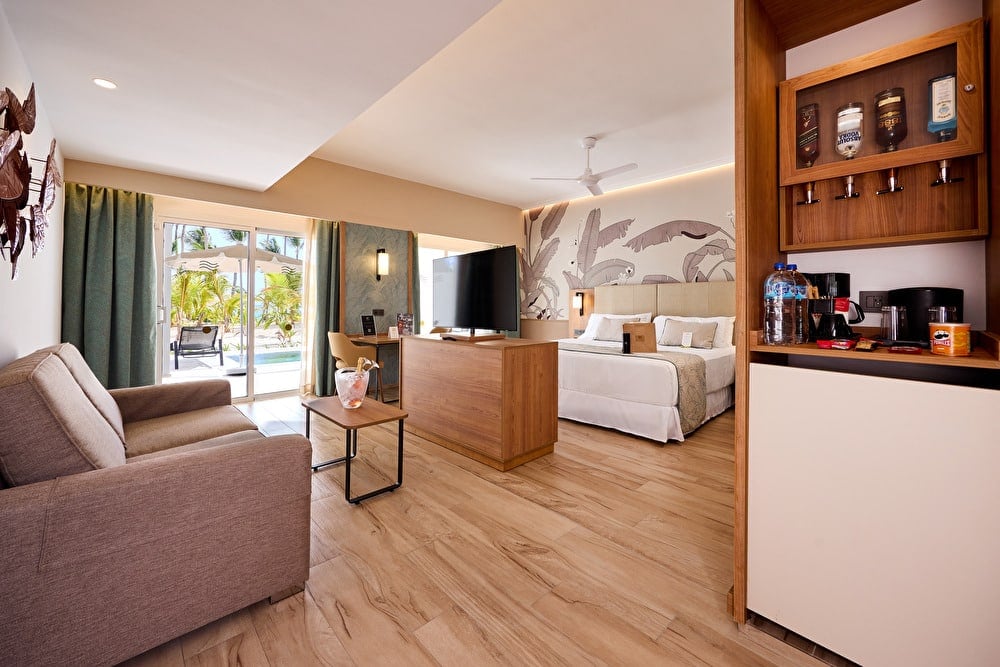 Vista general de una amplia suite de hotel con zona de estar, cama King, dispensador de licores en el mueble minibar y acceso a terraza exterior.