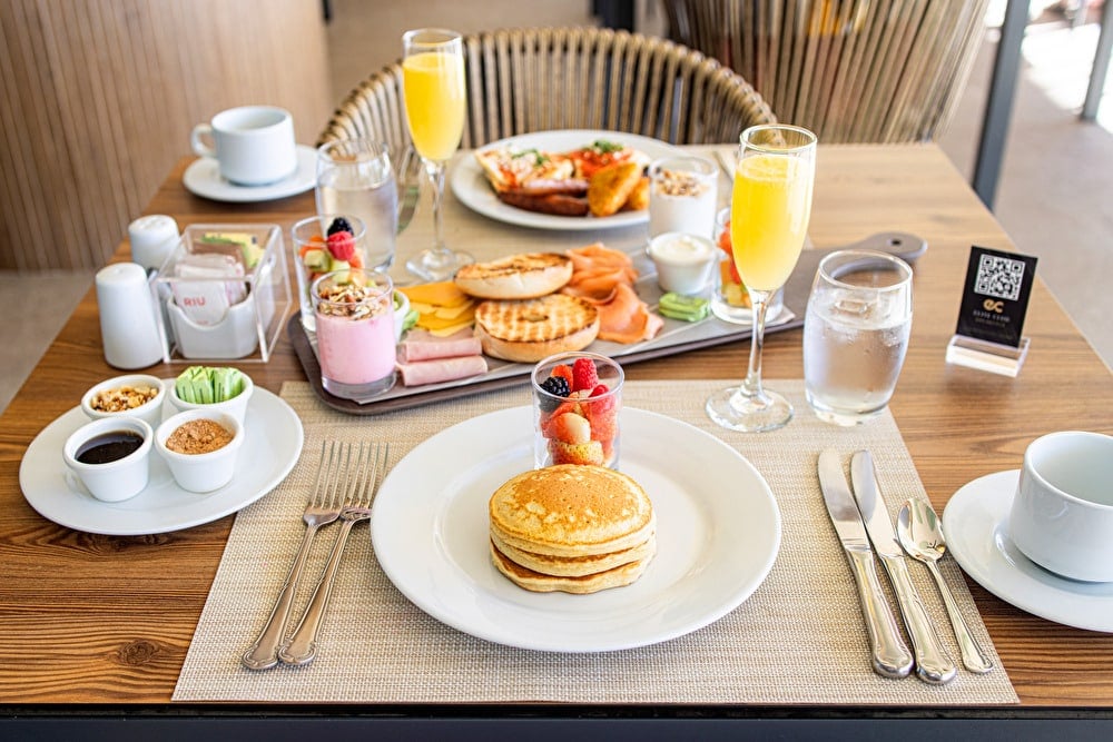 Mesa servida con un desayuno completo que incluye pancakes, bagels con salmón, yogur, fruta fresca y copas de mimosa en el área de Elite Club.