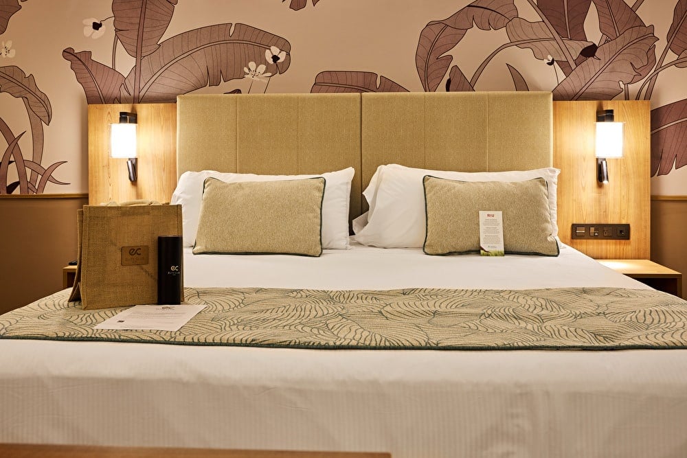 Cama doble de hotel preparada con detalles de bienvenida de Elite Club y pared decorada con papel tapiz de hojas tropicales.