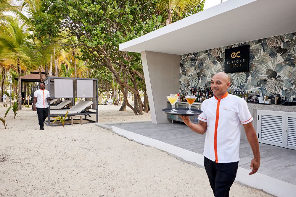 Camareno sirviendo cócteles en bandeja junto al bar de playa de Elite Club con camas balinesas al fondo.