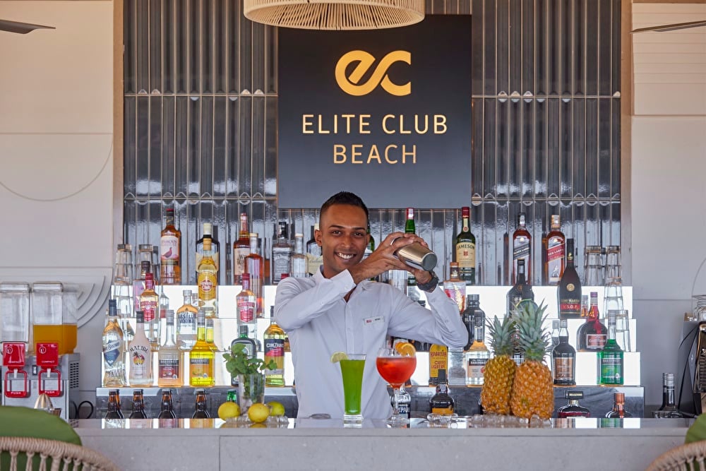 Bartender preparando un cóctel en la coctelera tras la barra del Elite Club Beach con botellas de licores premium al fondo.