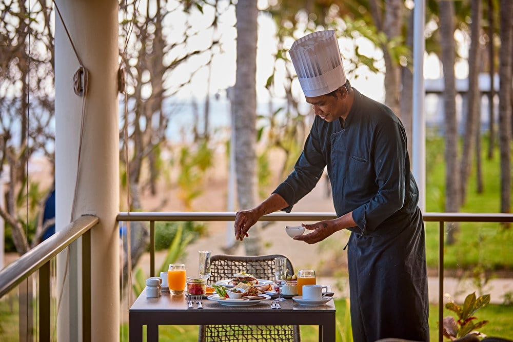 Chef emplatando un desayuno gourmet en una mesa de terraza al aire libre rodeada de vegetación tropical.