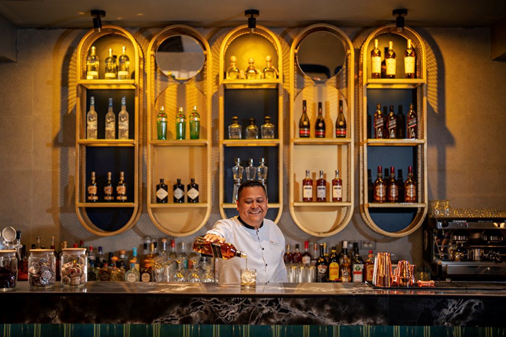 Bartender sirviendo un trago en la barra de un salón lounge con estanterías retroiluminadas llenas de botellas de licor.