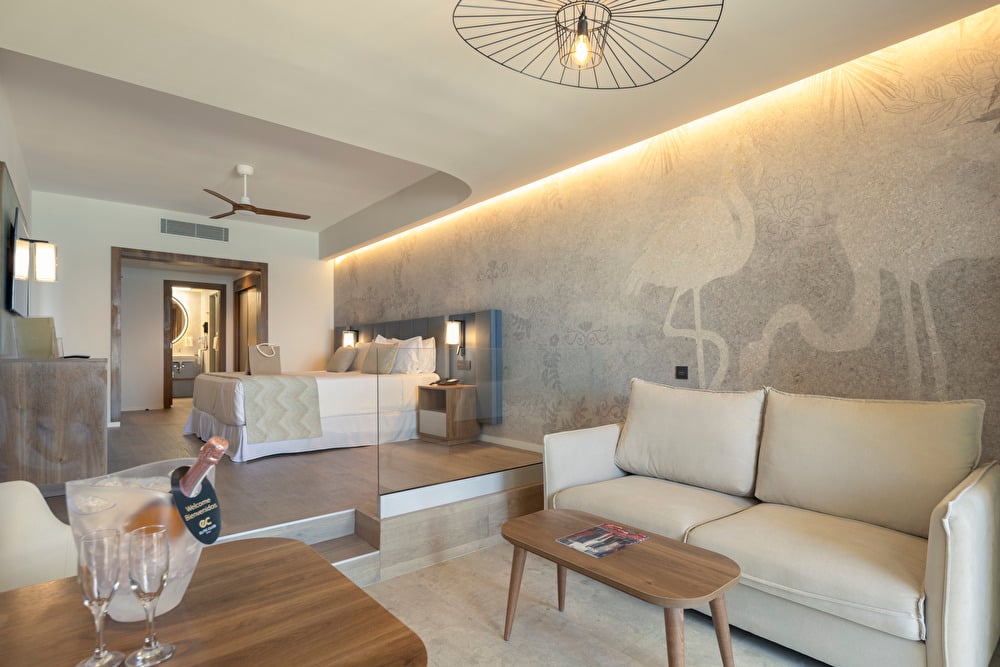 Junior Suite at Riu Palace Aruba featuring a double bed and a seating area with a sofa, decorated in cream tones with wooden furniture accents.