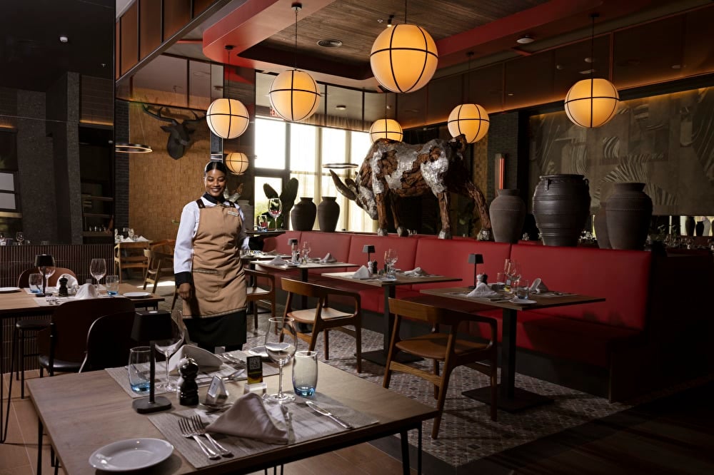 Steakhouse specialty restaurant at Riu Palace Aruba, featuring burgundy decor and soft, warm lighting, with a smiling waiter serving a table on the right.