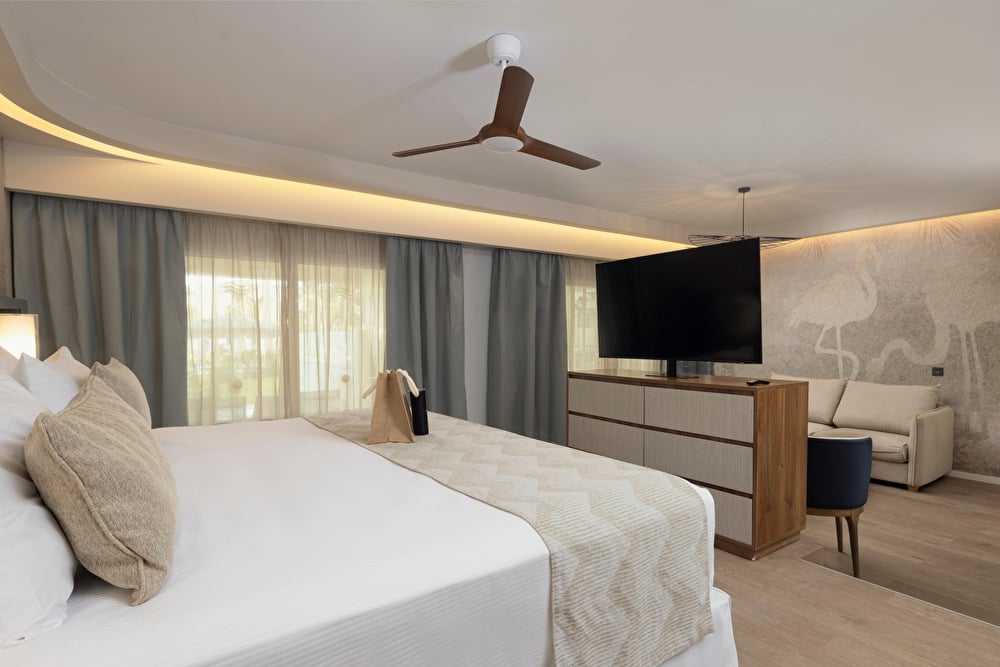 Suite at Riu Palace Aruba featuring warm, minimalist decor with cream tones and wooden accents, showing a bed facing a TV with a sofa positioned behind it.