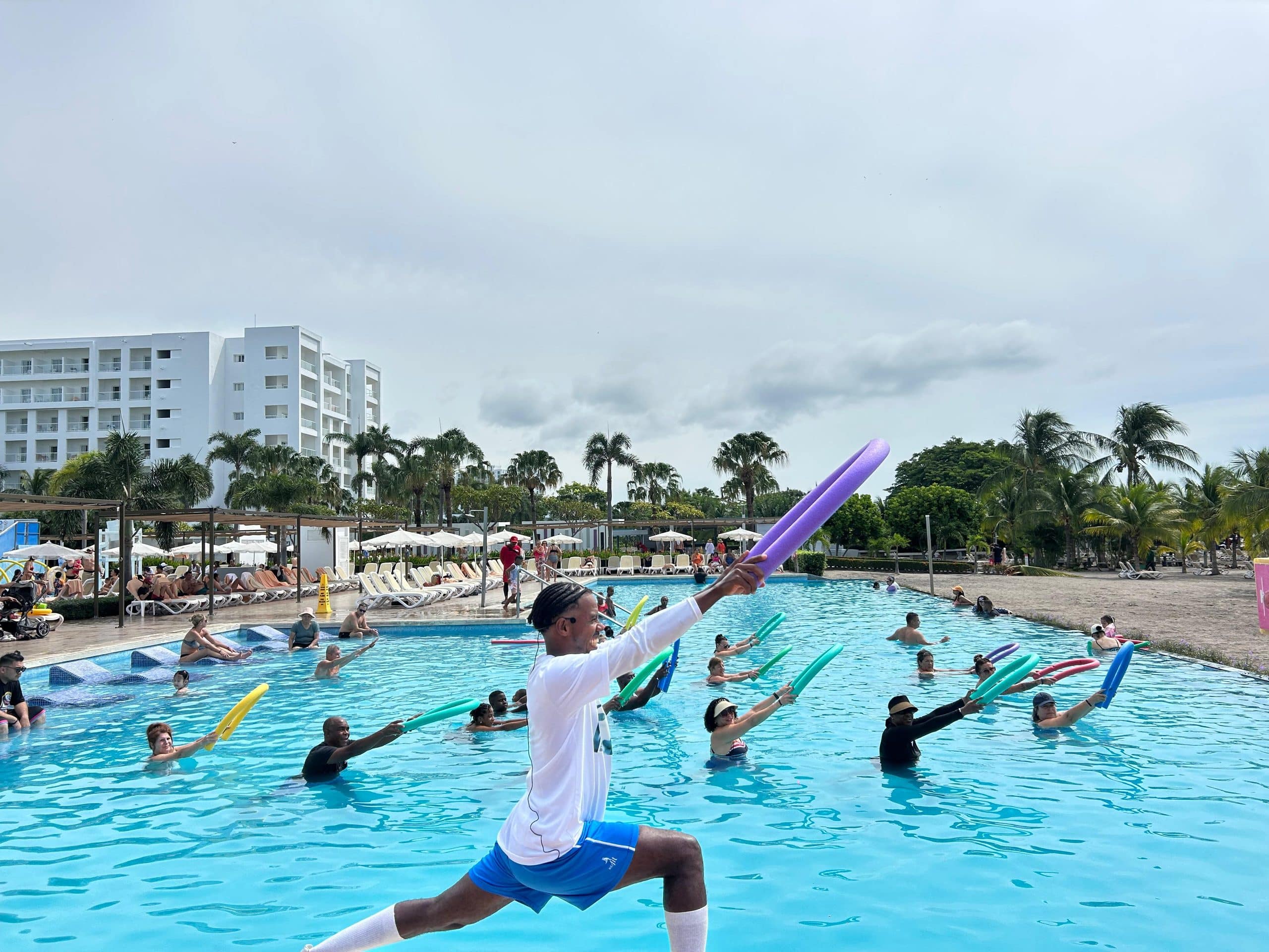 Un monitor de fitness con uniforme blanco y azul dirige una clase de aquaeróbic en una gran piscina de hotel, sosteniendo un churro de piscina morado.