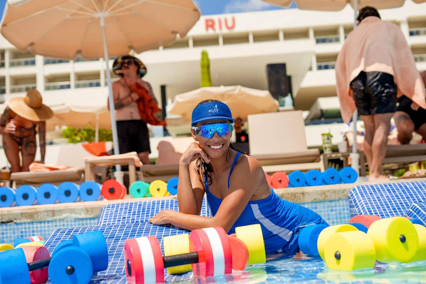 Monitora de animación RIUfit con visera azul posando junto a materiales acuáticos en la piscina del Hotel RIU Palace Baja California