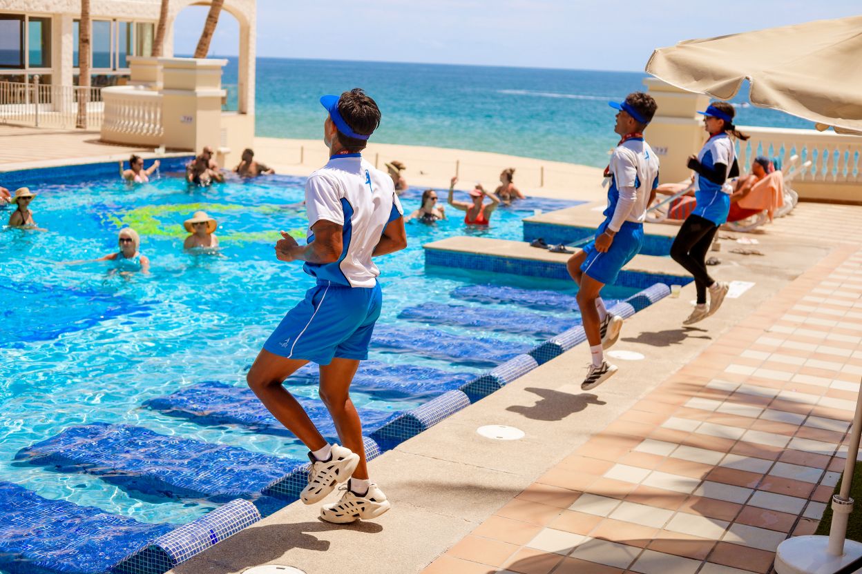 Equipo de animadores de RIUfit dirigiendo una clase de aquagym al borde de la piscina frente al mar en el Hotel RIU Palace Cabo San Lucas.