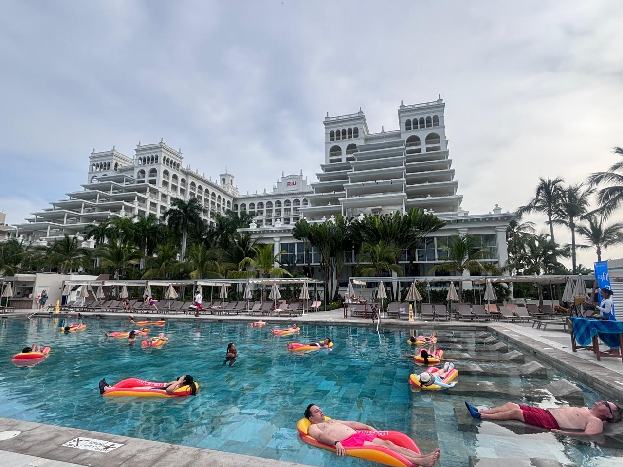 Un grupo de personas practica estiramientos sobre colchonetas de yoga en una terraza cubierta del hotel, con palmeras y la playa de fondo bajo la luz del sol.
