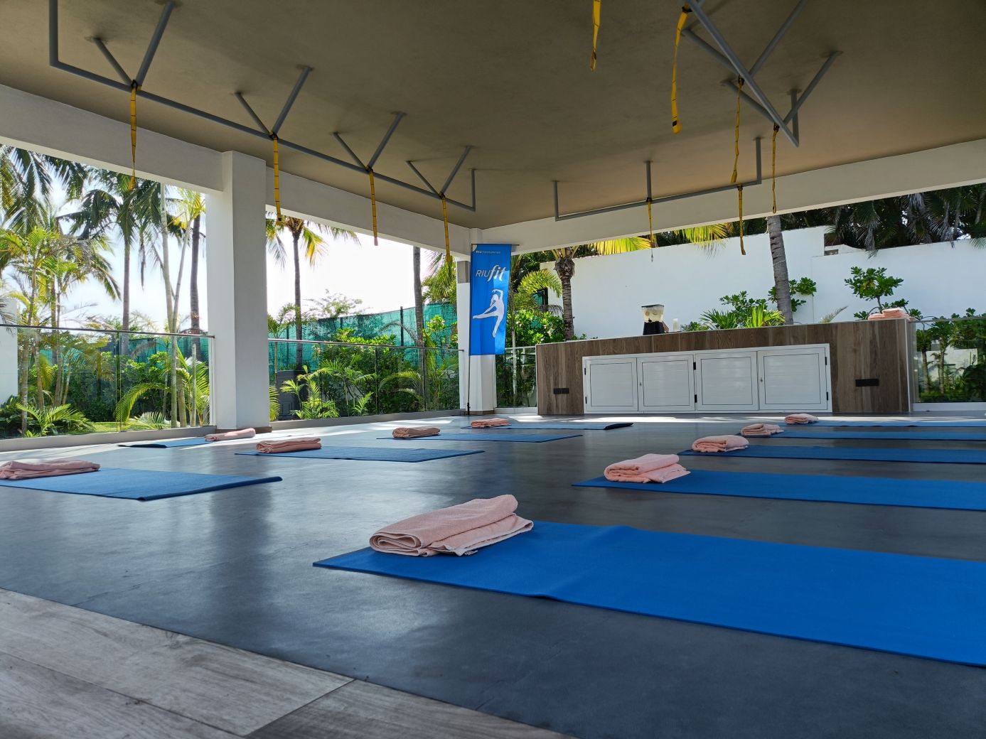 Esterillas azules con toallas rosas preparadas para una clase de yoga RIUfit en una terraza cubierta rodeada de palmeras en el Hotel Riu Vallarta.
