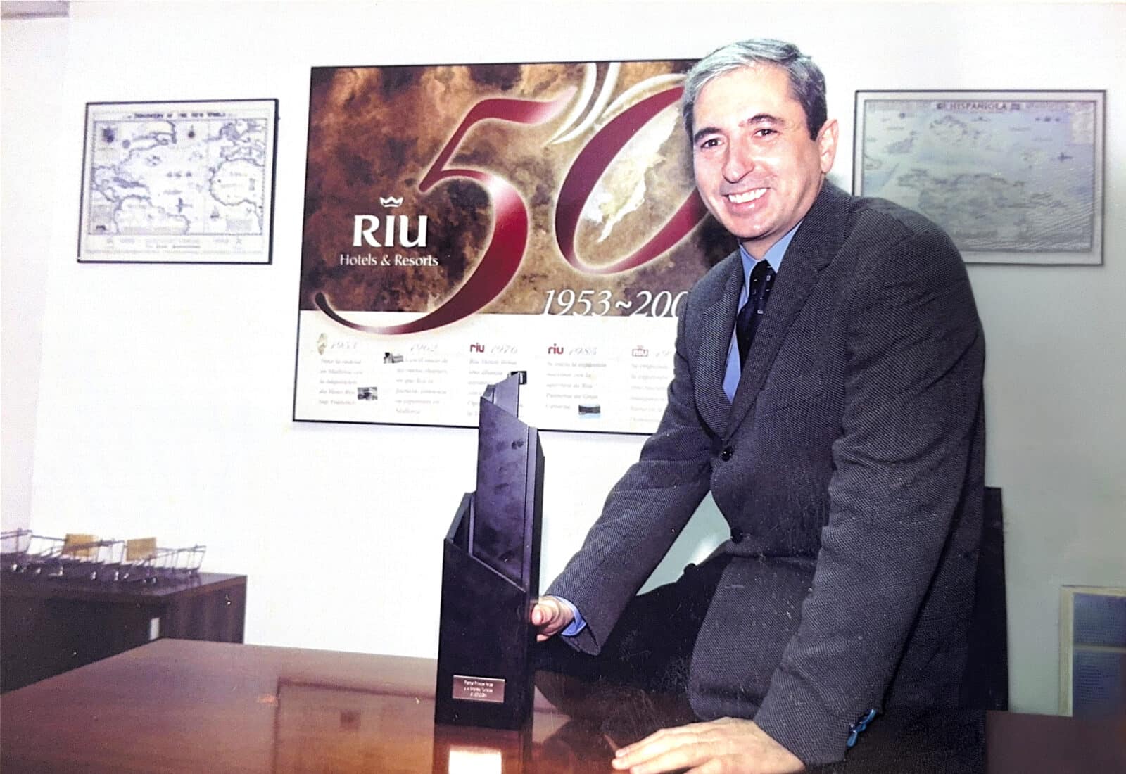 Fernando García en un despacho de RIU con un trofeo, ante el cartel del 50 aniversario de RIU Hotels & Resorts (1953-2003)