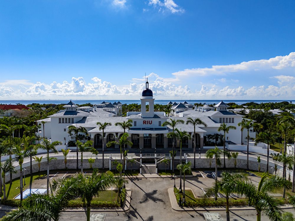 Vista aérea de la entrada principal del hotel Riu Palace Mexico, mostrando su fachada blanca de estilo colonial con una torre central, rodeada de palmeras y el mar de fondo.