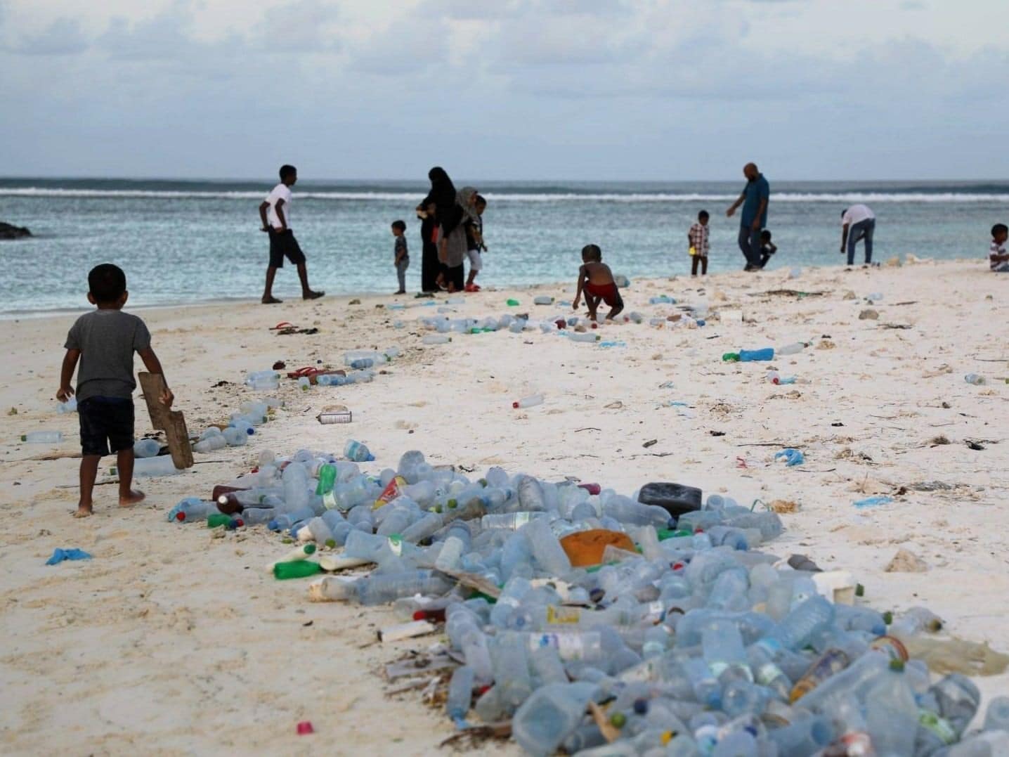 Foto de una playa tropical contaminada con una densa acumulación de botellas de plástico. Varias personas y niños están en la escena, evidenciando el impacto ambiental de los desechos plásticos cerca del océano.