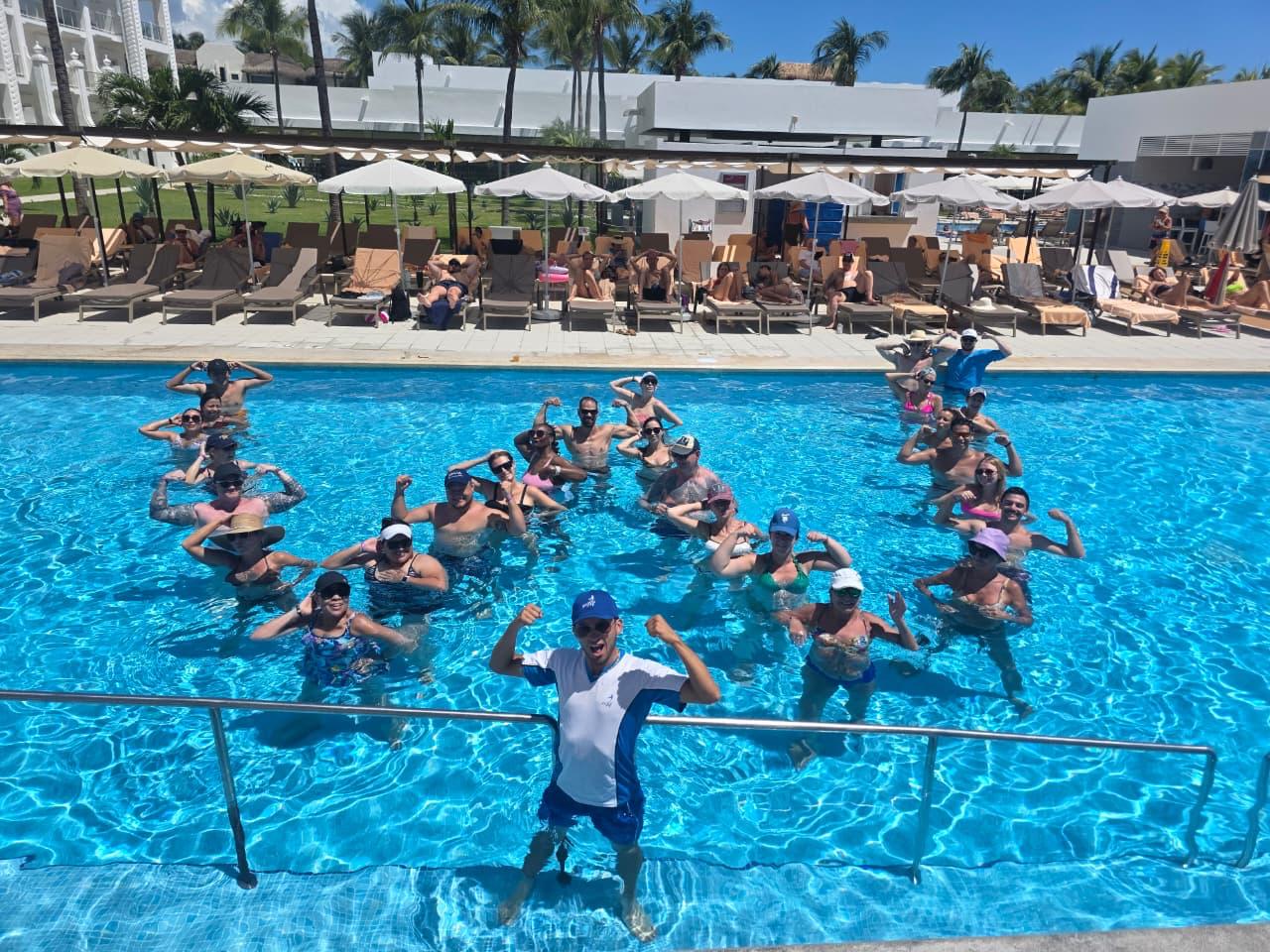 Vista general de huéspedes y monitor posando en la piscina con los brazos flexionados formando la letra W durante una sesión de aquagym en el Hotel Riu Palace Riviera Maya