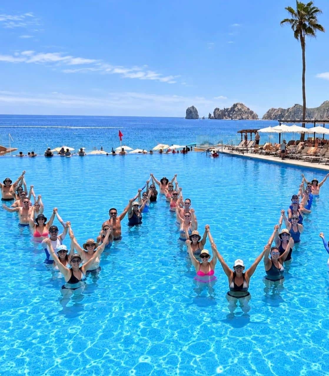 Huéspedes en la piscina infinita con vistas al mar formando la letra W tomada de las manos durante el World Wellness Weekend en el Hotel Riu Santa Fe