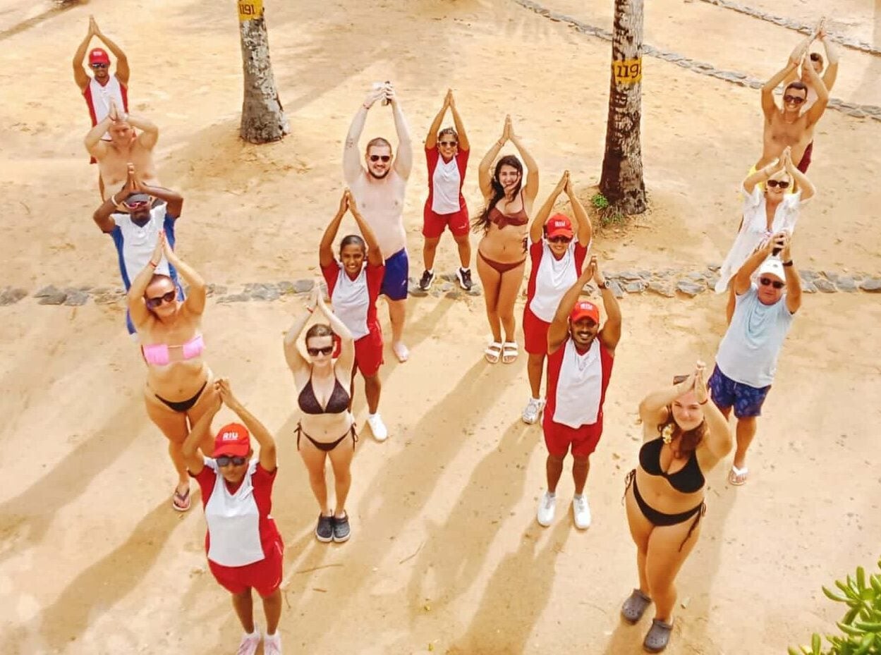 Vista cenital de huéspedes y equipo de animación formando la letra W con los brazos unidos sobre la cabeza durante una sesión wellness en el Hotel Riu Sri Lanka