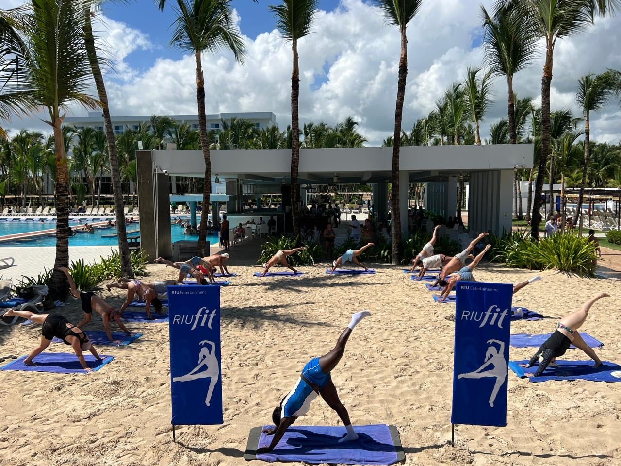 Grupo de personas realizando la postura del perro boca abajo con elevación de pierna sobre la arena durante una clase de yoga RIUfit en el Hotel Riu Republica