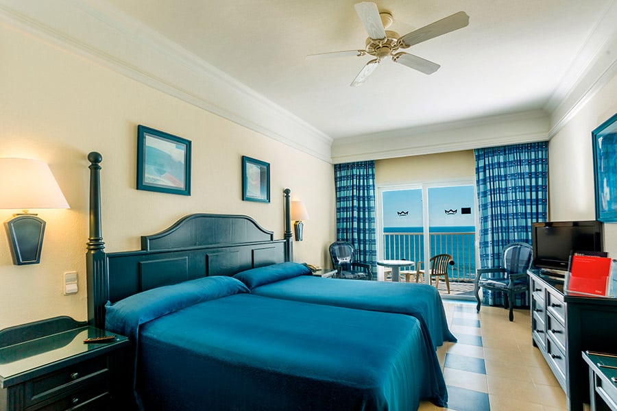 Hotel room with two queen beds, blue bedding, balcony access, ceiling fan.
